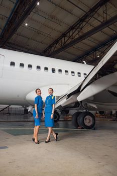 Full length shot of two air stewardesses in stylish blue uniform smiling at camera, posing together in front of passenger aircraft in hangar at the airport. Occupation concept