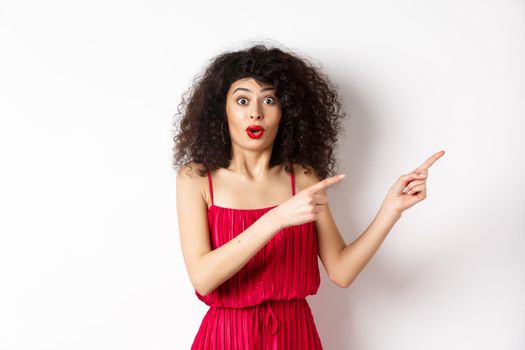 Surprised woman in red dress and makeup pointing fingers right, showing logo and look intrigued, standing on white background.