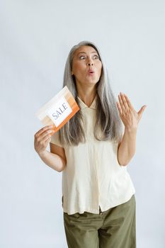 Emotional hoary haired Asian woman in casual clothes holds colorful card with word Sale on light grey background in studio
