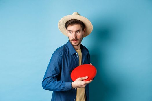 Handsome young guy in summer hat playing frisbee, throwing it aside, standing on blue background.