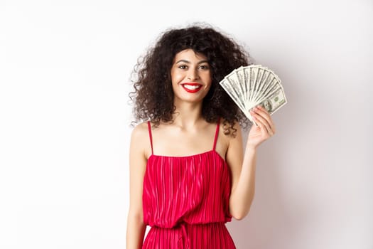 Stylish smiling woman in red dress showing money, standing on white background.