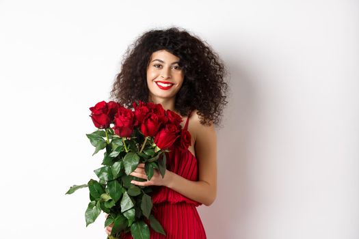 Gorgeous caucasian woman in red evening dress receiving bouquet of flowers. Girl with red roses smiling at camera, having romantic date on Valentines lover day, white background.