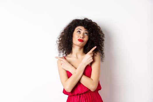 Indecisive silly woman in red dress, cross fingers and pointing sideways, looking up pensive, making choice, standing over white background.