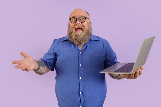Joyful middle aged fat businessman in tight blue shirt with spectacles holds laptop posing on purple background in studio