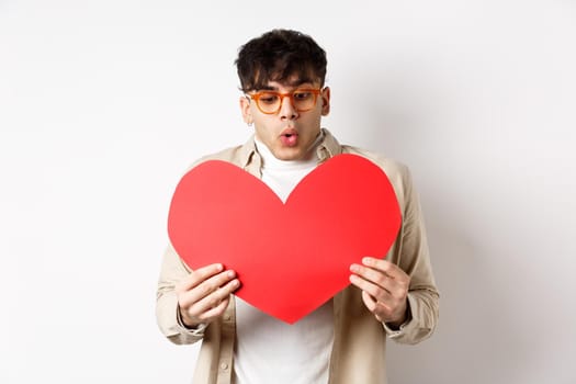 Surprised handsome man receive big red heart postcard on Valentines day, looking at gift with amazement, enjoying lovers day, standing over white background.