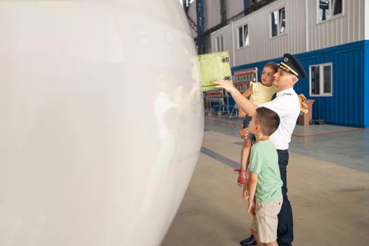 Professional pilot in uniform showing parts of airplane to his two little kids who came to visit their father at the aircraft hangar. Aircraft, family, childhood concept