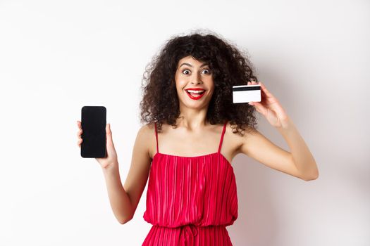 Online shopping concept. Excited curly-haired woman in red dress showing empty smartphone screen and plastic credit card, smiling happy at camera, standing on white background.