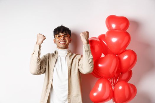 Valentines day. Satisfied young man saying yes, triumphing and celebrating on lovers date, making fist pump and smiling pleased, standing near heart balloons on white background.