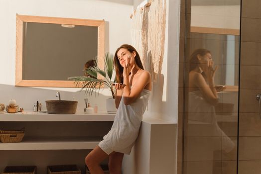 A young woman applies moisturizer to her face in her sun bath. Daily routine, hydration and skin care concept.