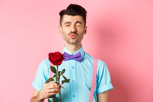Cute and funny man waiting for kiss from lover on Valentines day, holding beautiful red rose for girlfriend, standing over pink background.