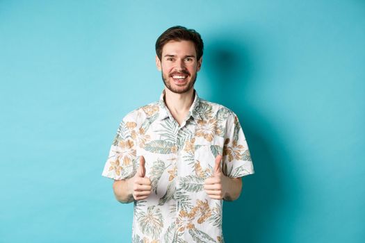 Summer holiday. Happy tourist showing thumbs up in approval and smiling, wearing hawaiian shirt, recommending travel agency, blue background.