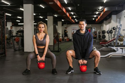 Fitness couple in sportswear squatting with weights and doing squats in the gym. A personal trainer in the gym corrects the squats of a young athlete.