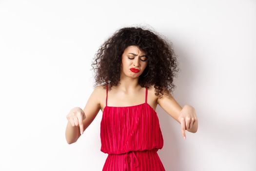Sad and disappointed curly-haired woman in red dress, frowning and looking down jealous, pointing at promo, standing over white background.