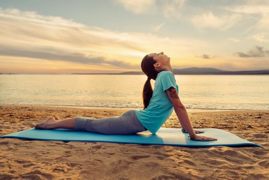 Sporty young woman stretching her back on yoga mat on sand beach near the sea in summer outdoor.