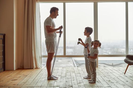 Father with resistance bands is teaching happy daughter and son exercising with dumbbells before window
