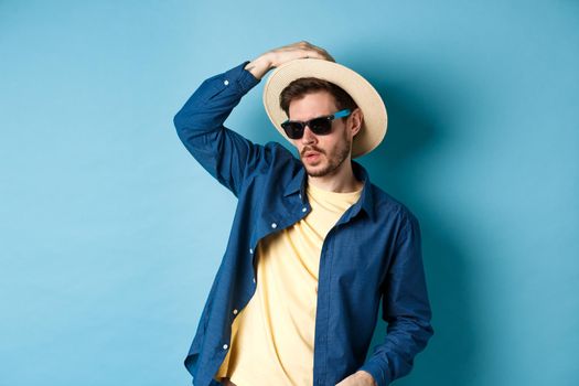 Cool guy having fun on vacation, wearing straw hat and sunglasses, looking aside sassy, standing on blue background.