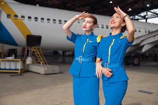 Two air stewardesses in stylish blue uniform smiling while looking up in the sky, standing together in front of passenger aircraft in hangar at the airport. Occupation concept