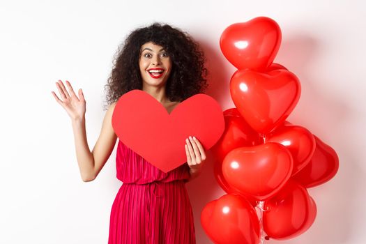 Valentines day and love concept. Cheerful young woman in elegant red dress, standing near romantic balloons and holding big red heart cutout, waving hand to say hi, waiting for date.