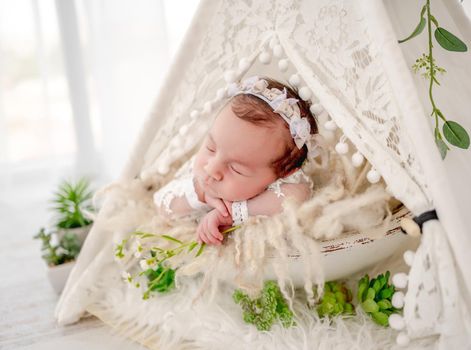 Adorable newborn baby girl wearing beautiful dress and wreath lying in hut wigwam with plant decoration holding hands under her cheeks in studio. Cute infant child napping on fur closeup portrait
