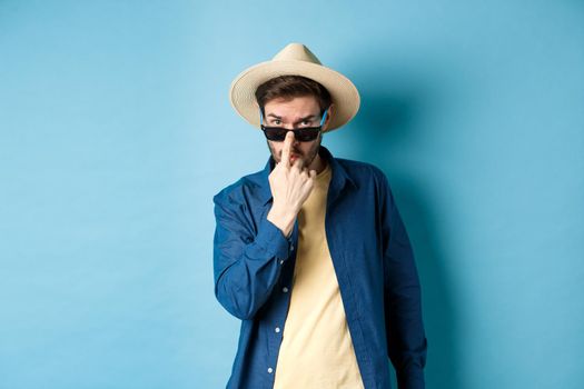 Handsome and cheeky guy put on sunglasses and straw hat, standing ready for summer vacation or tropical holiday, blue background.