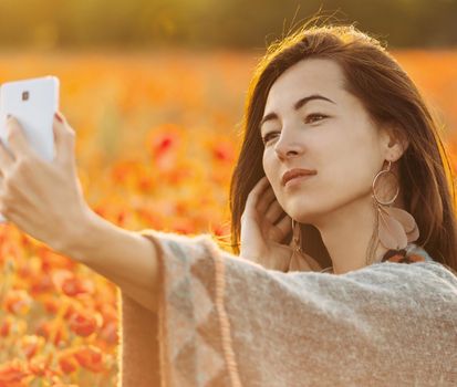 Attractive young woman boho stylish taking selfie portrait with smartphone in poppies flowers meadow at sunset.