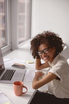 Smiling pretty lady in glasses sitting at workplace while using laptop