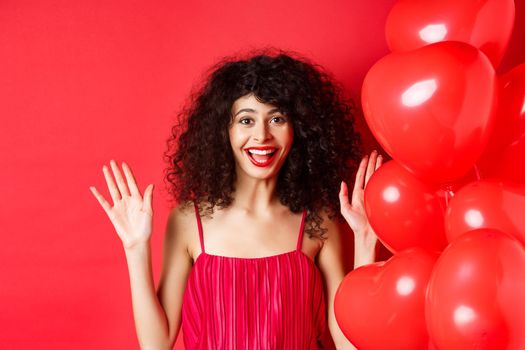 Excited stylish woman with curly hair, wearing dress, raising hands up and laughing happy, standing near heart balloons, white background.