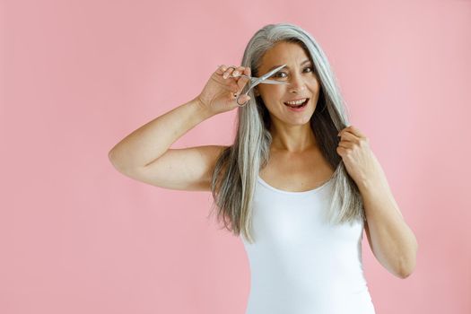 Happy middle aged Asian woman with long hoary hair looks through open scissors on pink background in studio. Mature beauty lifestyle