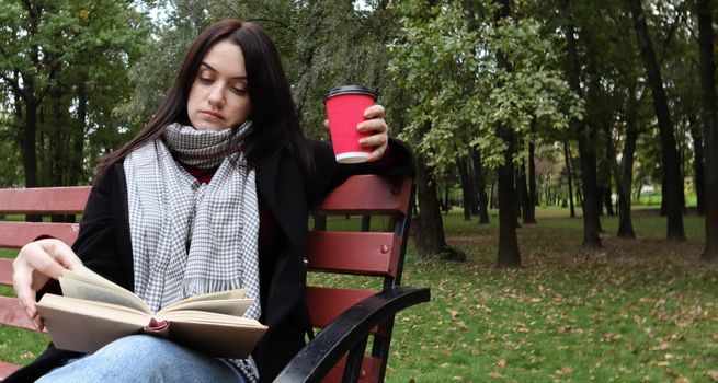 Young woman in jeans, coat and scarf, on a park bench. A woman is reading a book and drinking coffee or other hot drink outdoors alone
