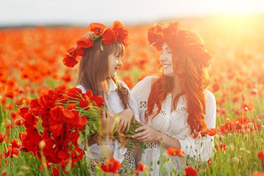 Little girl with redhead mother in white dresses and wreathes poses with bouquet of poppies on poppy field at warm summer sunset