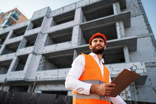 Young man engineer in workwear standing in construction site with clipboard, close up portrait