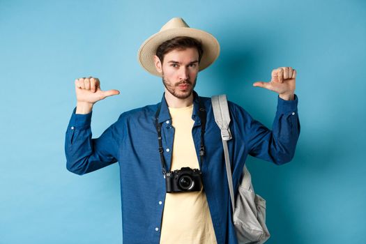 Confident handsome guy in summer hat, pointing at himself with bragging look, going to travel on holidays, holding backpack and camera, blue background.