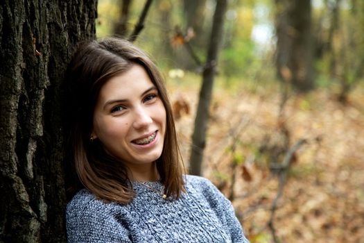 Young beautiful girl with braces on her teeth in a gray sweater sits in the autumn forest near a large tree and smiles sweetly.