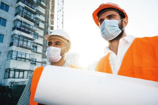 Two men in workwear and medical masks working with blueprints on object, close up