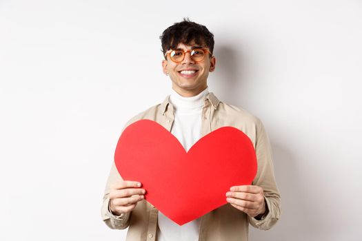 Attractive modern man smiling and looking at camera hopeful, holding big red Valentines heart, waiting for soulmate on lover day date, standing over white background.