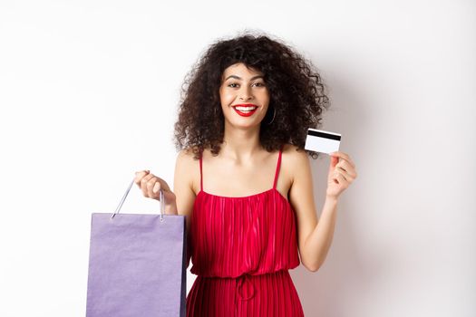 Beautiful female model with curly hair, red dress, showing shopping bag and plastic credit card, white background.