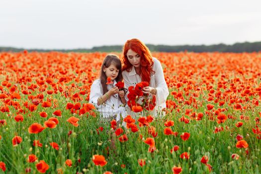 Little happy girl with redhead mother in white dresses makes wreath on poppy field at warm summer sunset