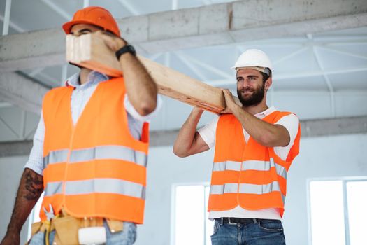 Two young men builders carrying wood planks on construction site, close up photo