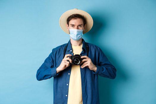 Covid-19, pandemic and travel concept. Cheerful tourist in summer hat and medical mask taking photos, holding camera, standing on blue background.