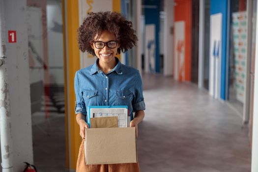 Happy Afro American woman in glasses standing in the hallway while holding paper box, copy space