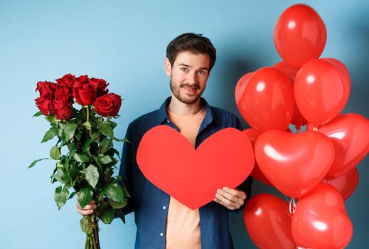 Cute guy waiting for girlfriend with valentines day presents, holding bouquet of roses and red heart, smiling at camera, standing over blue background.