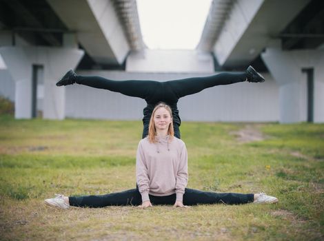Two girls acrobats perform stand in the splits on the grass against the background of the bridge. Wide shot