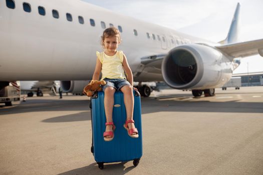 Full length shot of cute little girl holding her toy, looking at camera and sitting on suitcase in front of big airplane. Childhood, traveling, vacation concept