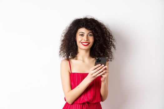 Cheerful woman with curly hair, using smartphone in red dress, smiling at camera, standing over white background. Copy space