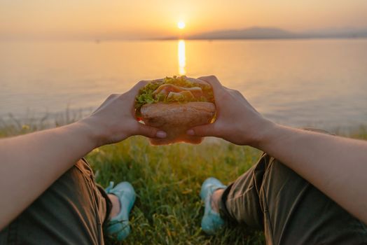 Woman resting by the sea with fresh tasty burger and enjoying view of summer sunset, point of view.