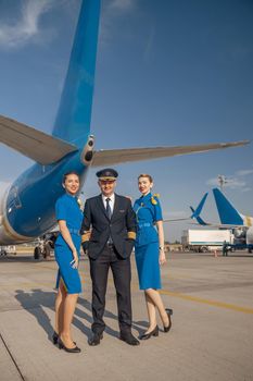 Full length shot of cheerful pilot and two pretty stewardesses standing together in front of an airplane and smiling at camera. Aircraft, aircrew, occupation concept