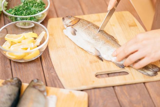 Female hands cutting raw trout fish on kitchen wooden board for seafood dish.