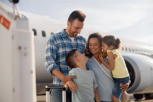 Family of four looking excited while going on a trip, standing in front of big airplane outdoors. People, traveling, vacation concept