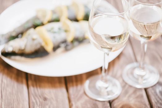 Baked fish dish with lemons and greenery and two glasses of white wine for couple on wooden table.