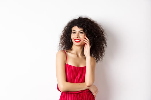 Elegant and romantic woman in red dress and makeup, smiling and touching her face, looking aside with happy expression, white background.